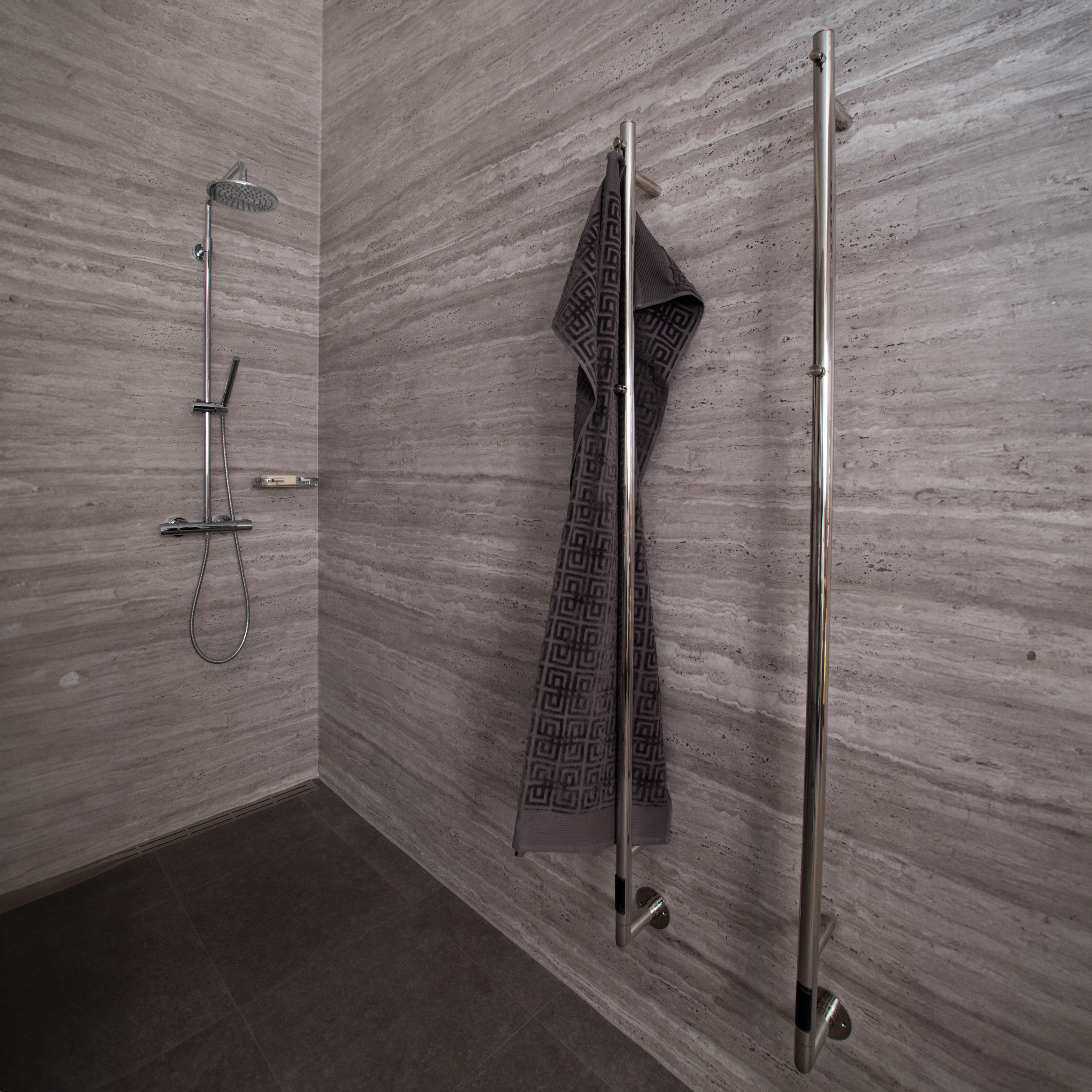 Ystad handdukstork, två stycken på en badrumsvägg. En handduk hänger på en av handdukstorkarna. Handdukstorkarna ser ut som pinnar med krokar i ovankant. I bakgrunden finns en dusch.