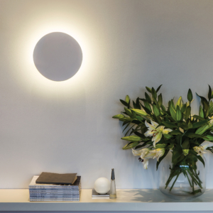 Eclipse Round 250 LED vägglampa sitter på en vägg vid ett bord med en bukett blommor och böcker.